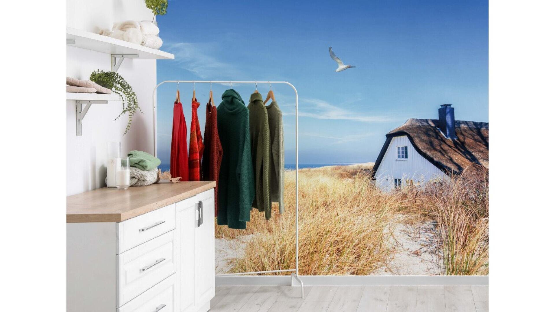 A bright, modern laundry room features a clothing rack with colorful sweaters. Behind it is a wall mural of a sandy beach, a cozy cottage with a thatched roof, tall grasses, and a seagull flying in a clear blue sky.