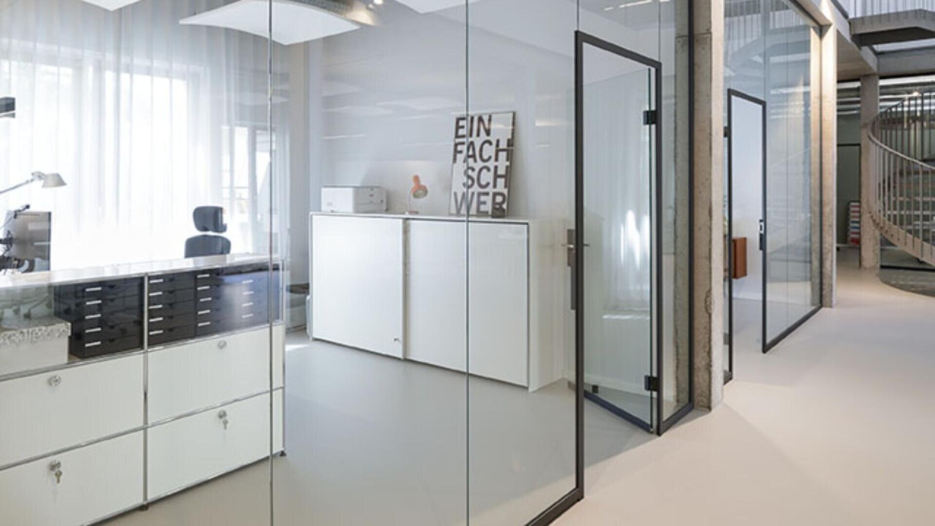 Modern office with glass walls, white cabinets, and a black office chair by a desk. A framed print with German text leans against the wall. Natural light filters through sheer curtains. A spiral staircase is visible in the background.