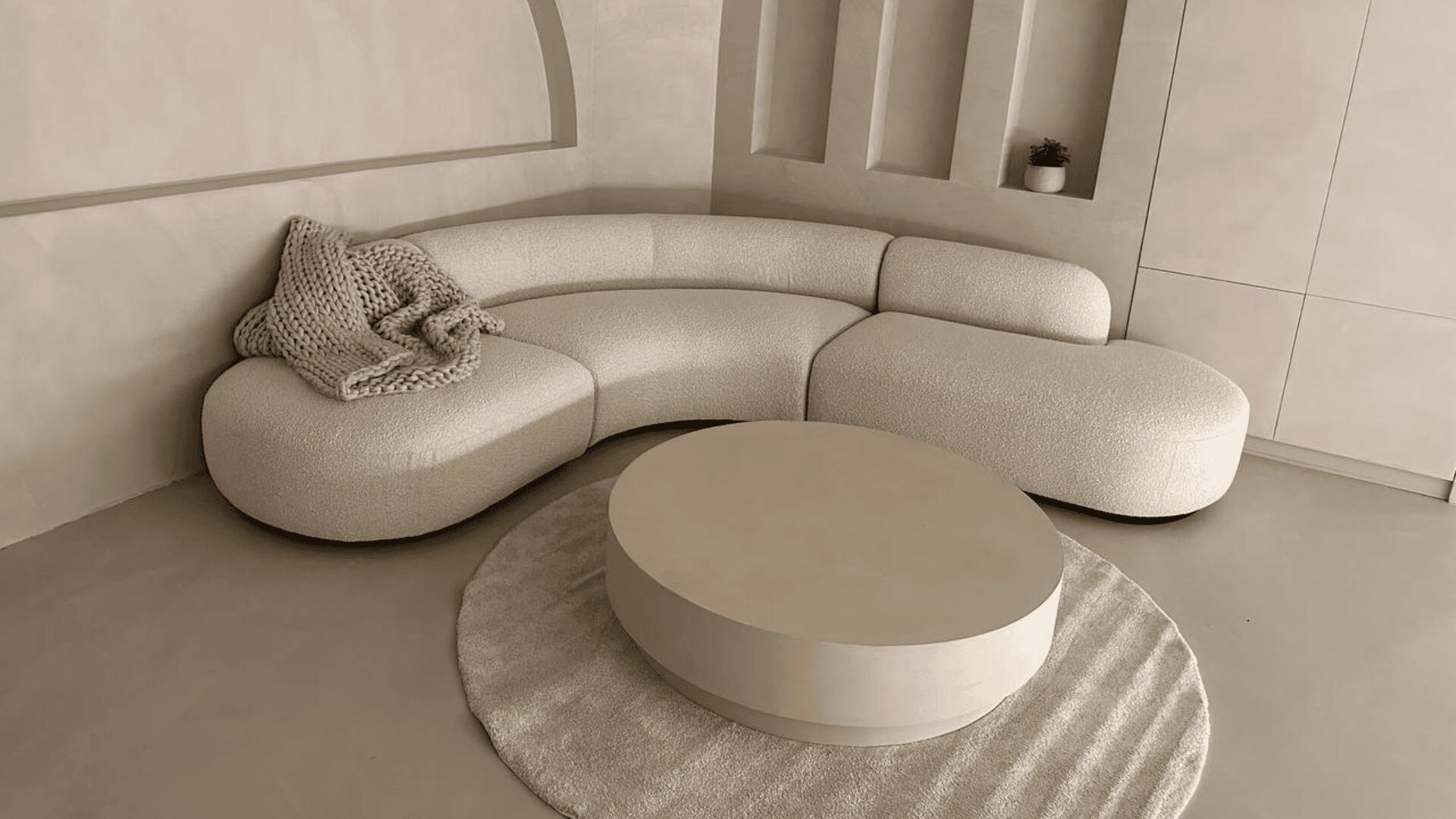 Ein modernes, geschwungenes weißes Sofa mit einer dicken Strickdecke steht vor einer beigen Wand mit eingebauten Regalen und einer kleinen Pflanze. Ein runder Couchtisch steht auf einem passenden runden Teppich.