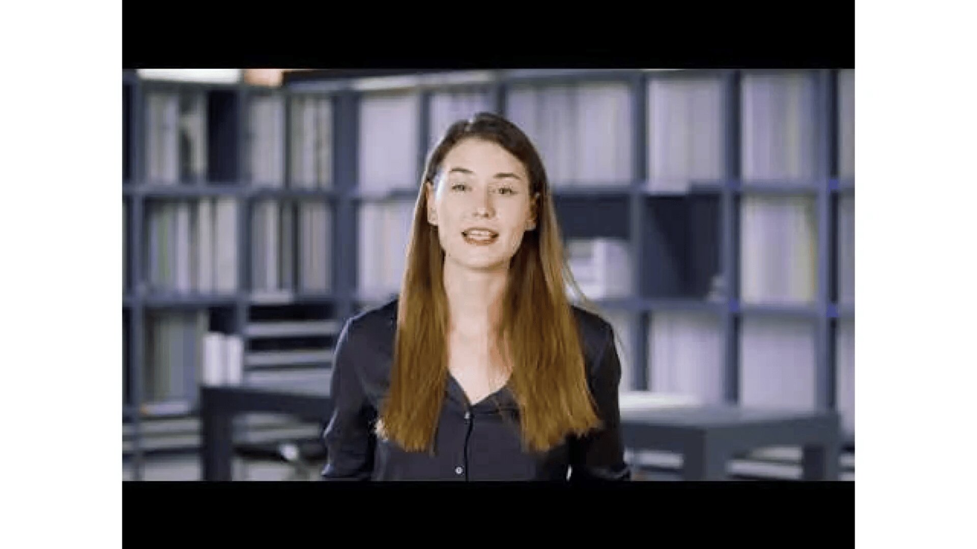 Eine Frau mit langen braunen Haaren und einem dunklen Hemd steht in einem Innenbereich vor Regalen voller Bücher oder Akten, blickt in die Kamera und spricht.