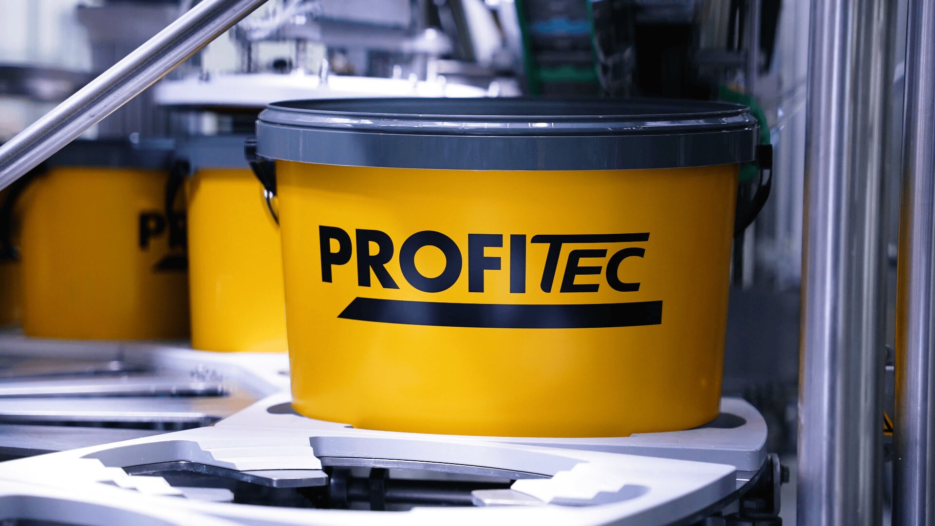Auf einer Industrieanlage steht ein leuchtend gelber Eimer mit der Aufschrift „PROFITEC“ in schwarzen Buchstaben, im Hintergrund sind Metall- und Kunststoffkomponenten zu sehen.