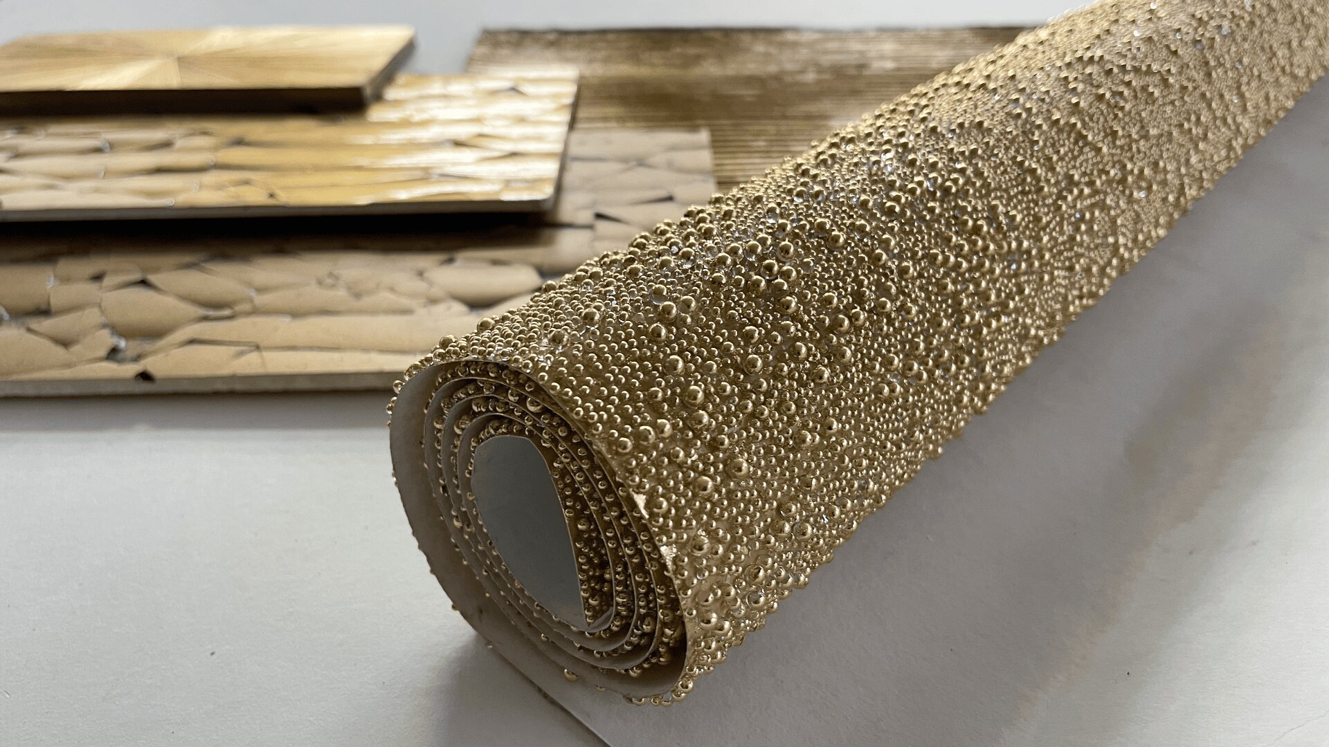 Auf einer weißen Fläche ist ein aufgerolltes Laken zu sehen, das mit kleinen Goldperlen bedeckt ist. Im Hintergrund sind mehrere strukturierte Goldplatten angeordnet.