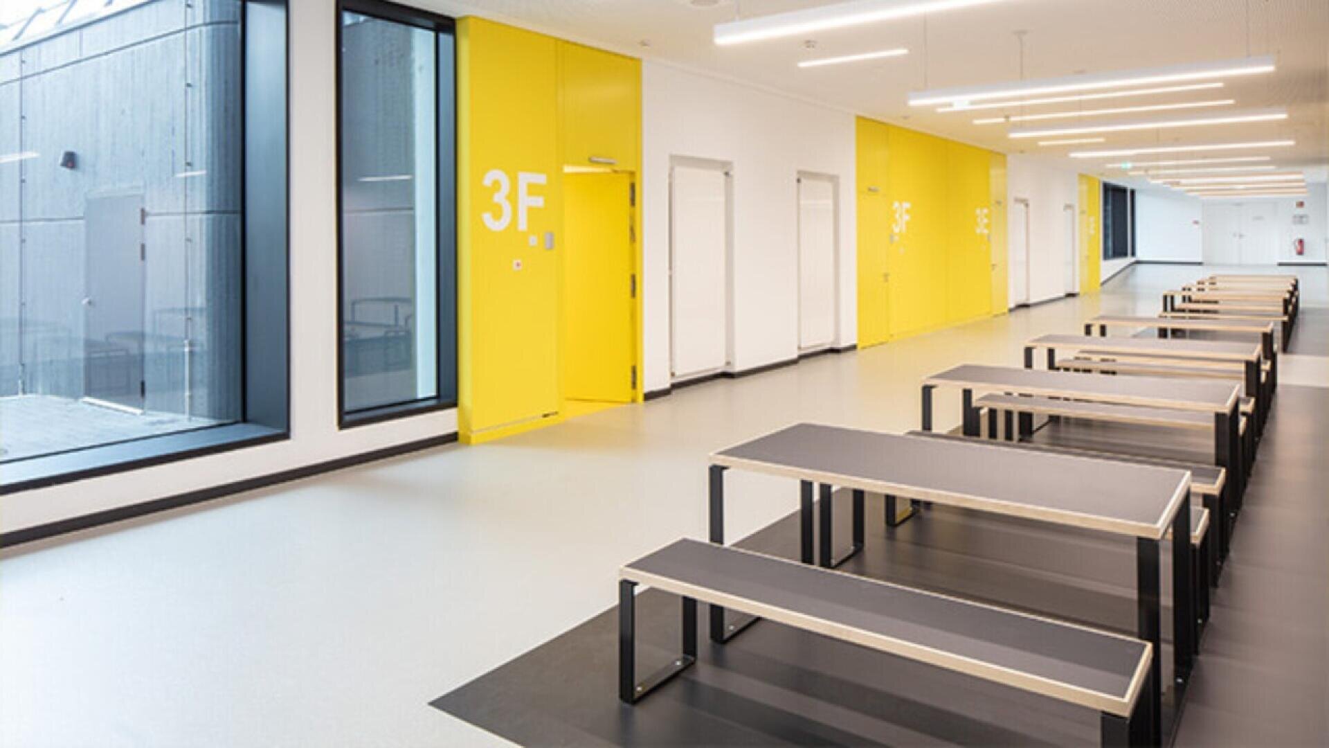 A modern, bright hallway with large windows, yellow doors labeled 3F, and several black tables with benches aligned along the wall. The space has minimalistic design and overhead lighting.