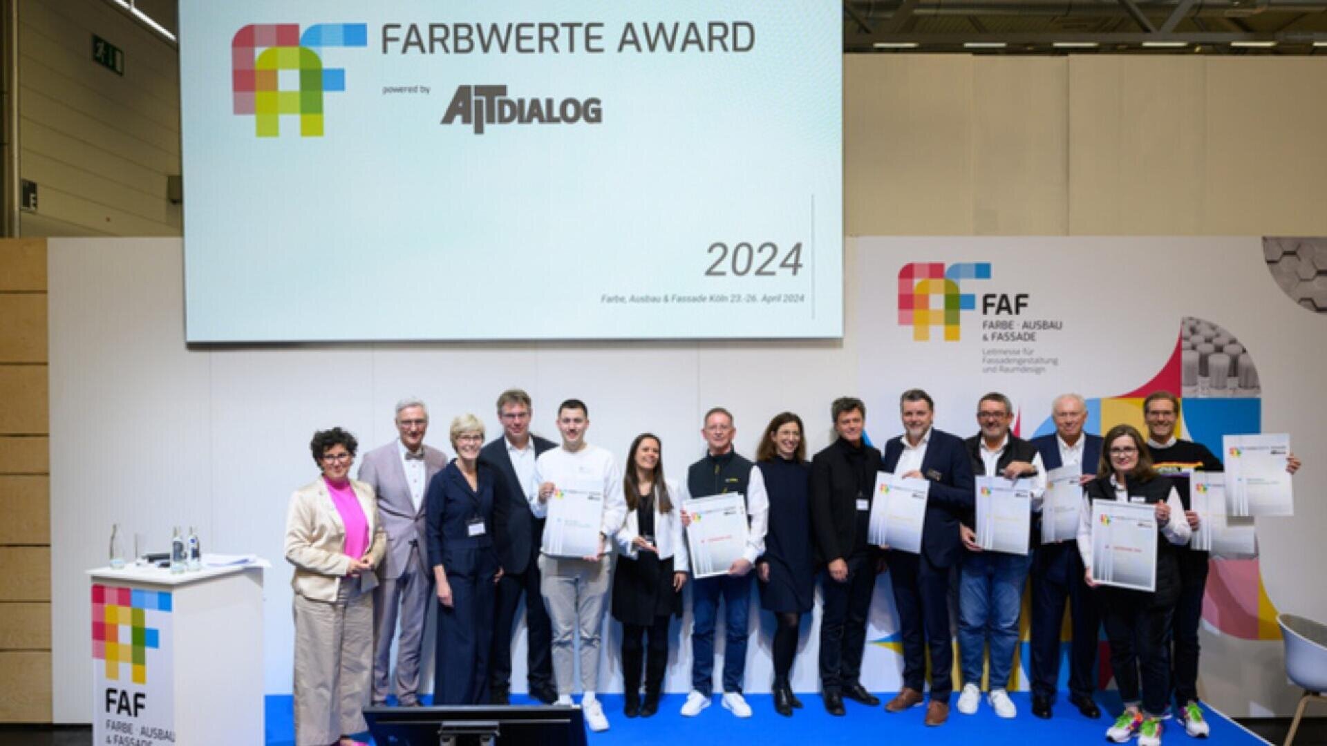 A group of people stands on a stage holding certificates at the Farbwerte Award 2024 event, with a large screen displaying the event name and logo in the background.