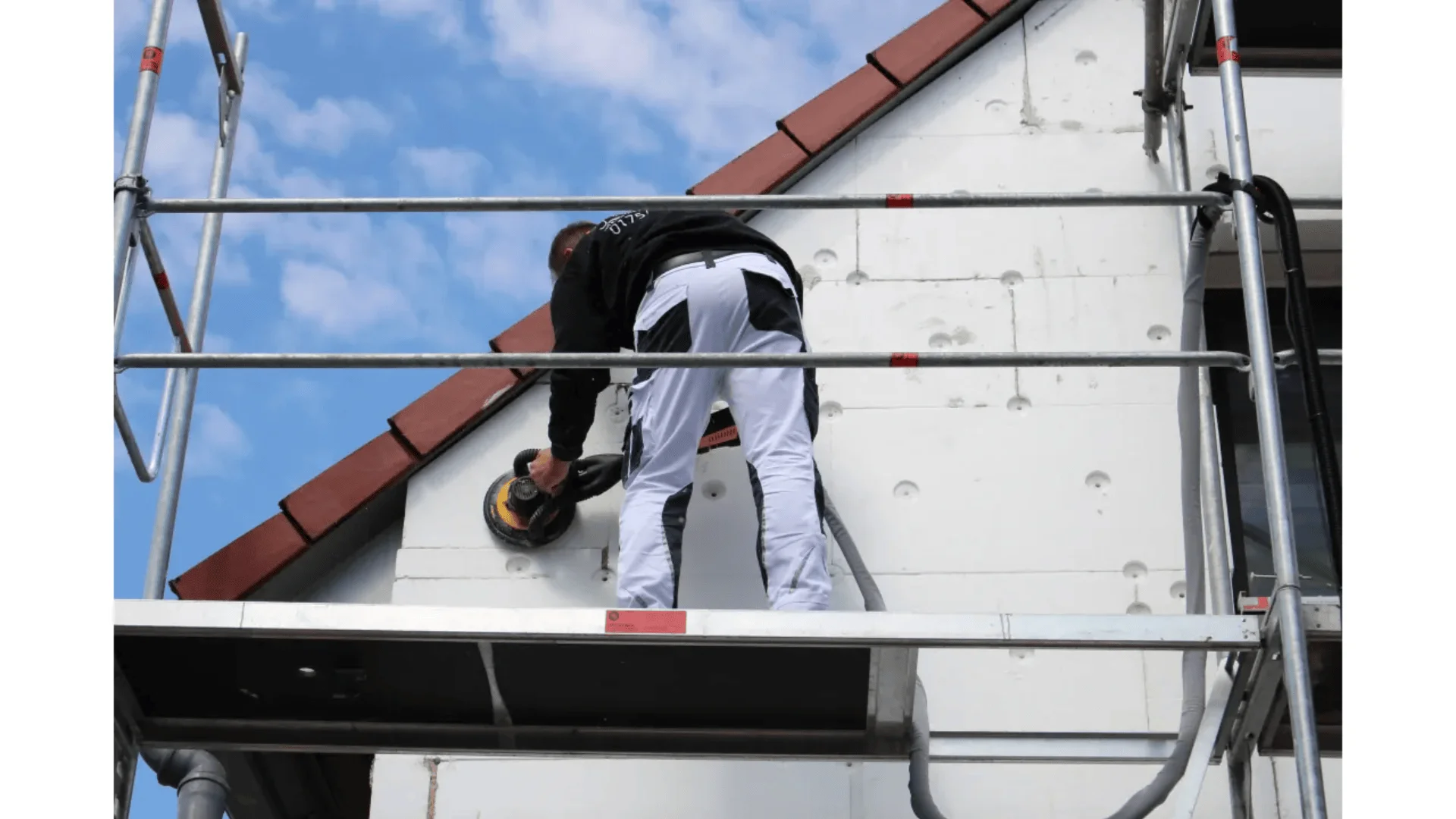Ein Arbeiter auf einem Gerüst schleift oder poliert die Außenwand eines Hauses, das gerade renoviert wird. Im Hintergrund sind blauer Himmel und Wolken zu sehen. Der Arbeiter trägt weiße Hosen und eine schwarze Jacke.