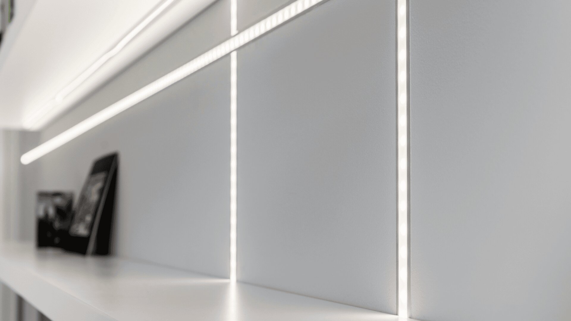 Nahaufnahme eines modernen weißen Regals mit minimalistischem Dekor, beleuchtet durch elegante, integrierte LED-Lichtstreifen, die horizontal und vertikal entlang der Wand verlaufen.