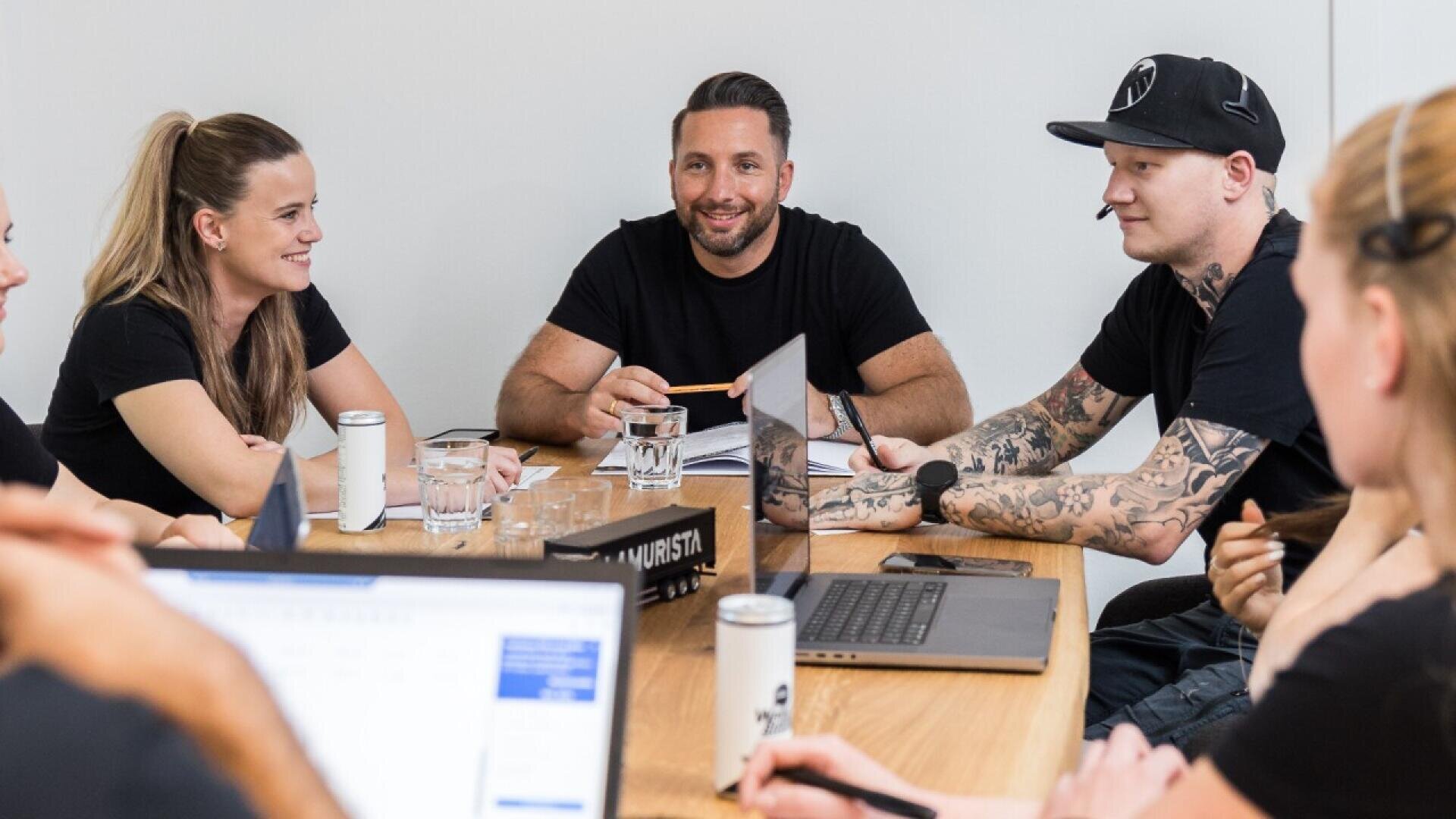 Five people sit around a table in a meeting room, talking and smiling. Laptops, drinks, and glasses of water are on the table. All are casually dressed and appear engaged in the discussion.