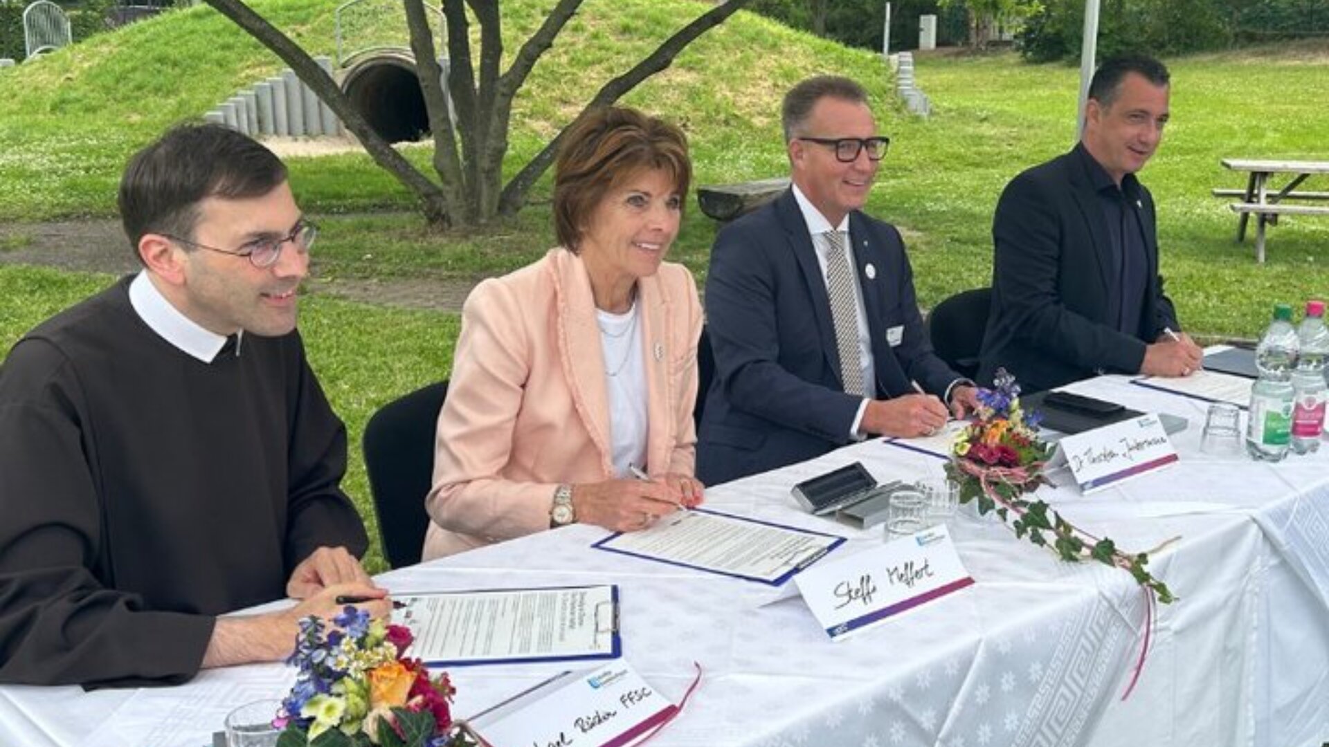 Vier Personen sitzen lächelnd an einem Tisch im Freien und unterschreiben Dokumente. Der Tisch ist mit einem weißen Tuch bedeckt und mit kleinen Blumenarrangements dekoriert. Im Hintergrund sind Bäume und Gras zu sehen.