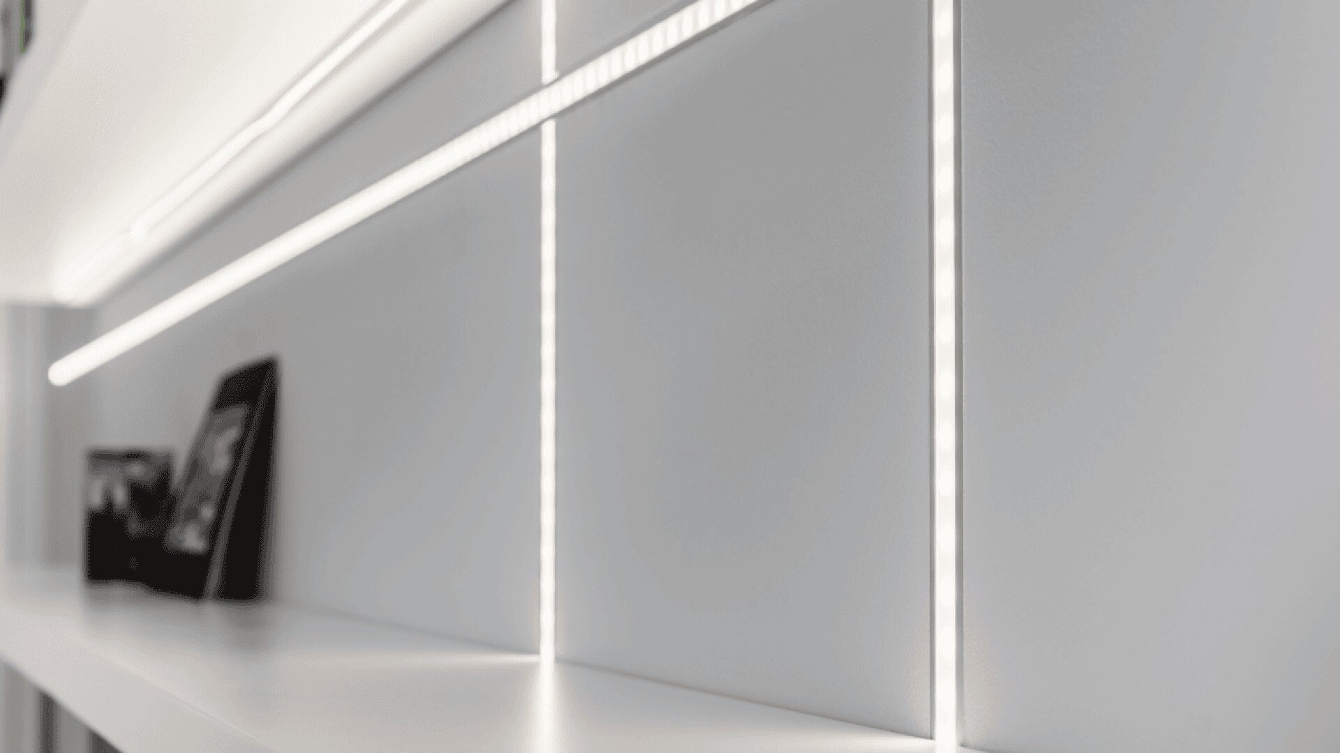A close-up view of minimalist white shelves with vertical and horizontal LED light strips, featuring a few blurred objects including a framed photo. The design creates a modern and clean aesthetic.