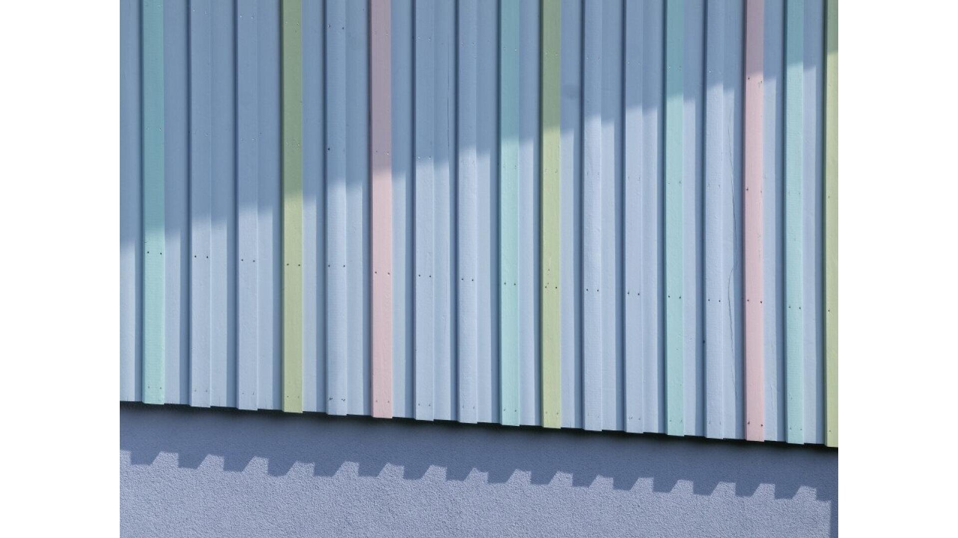 Corrugated metal wall painted in pastel blue, green, pink, and yellow vertical stripes, with a shadow cast diagonally across the surface and part of a blue base visible at the bottom.