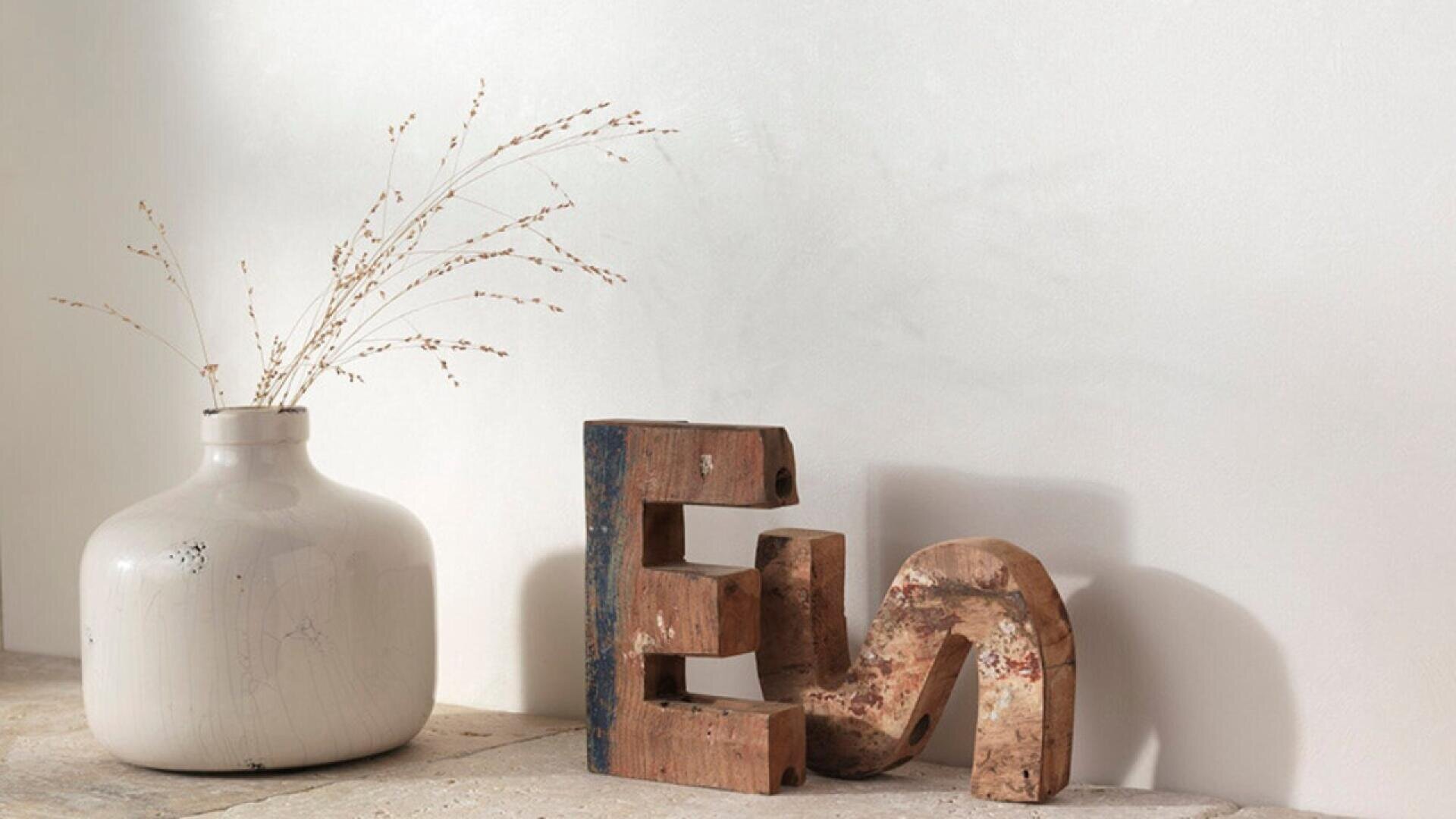 A white ceramic vase with dried grass stands next to two rustic wooden letter sculptures, “E” and “S,” on a light-colored stone surface against a plain, off-white wall.