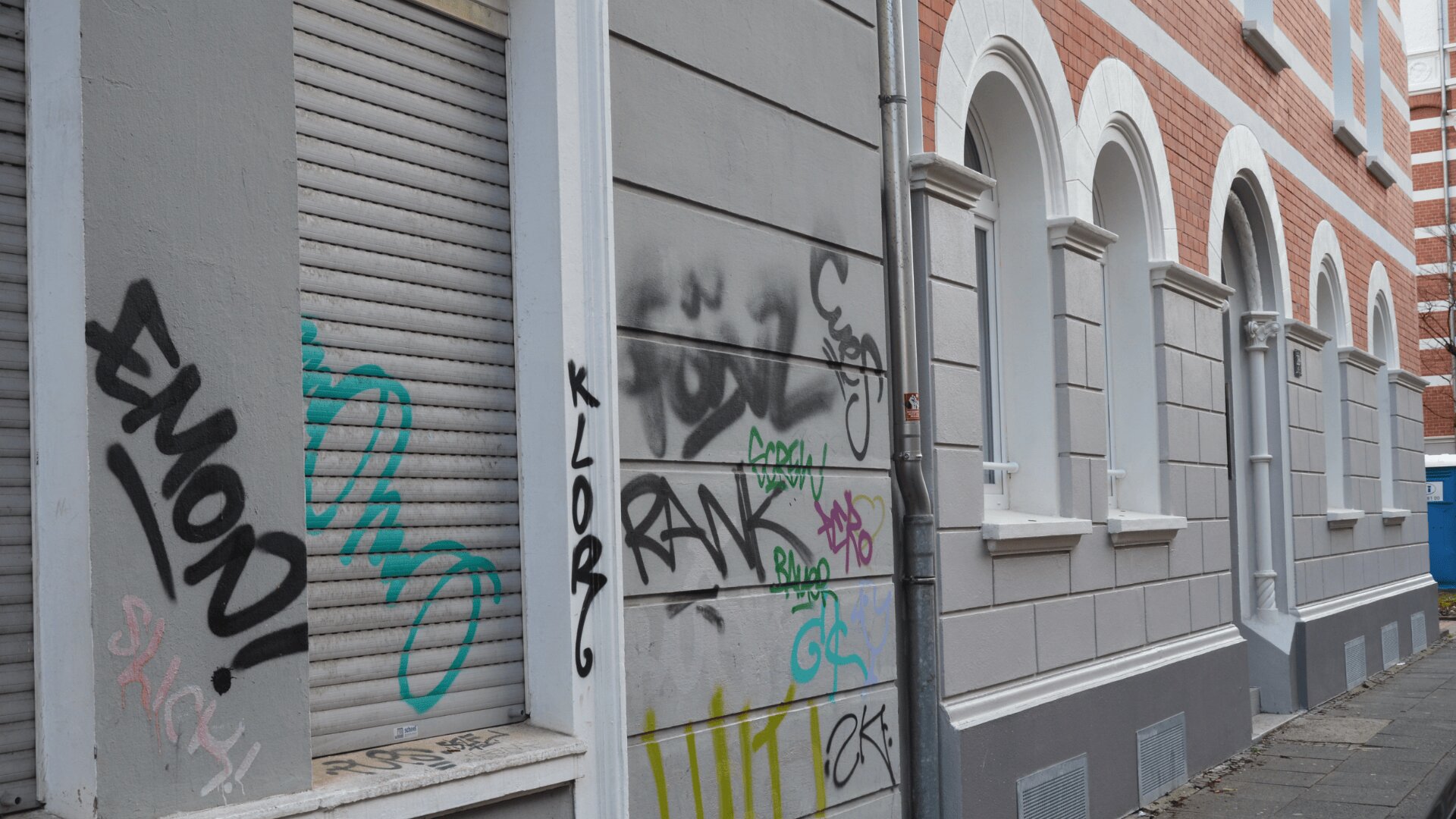 Ein Bürgersteig verläuft an einem Gebäude mit Bogenfenstern, teilweise geschlossenen Fensterläden und verschiedenen bunten Graffiti-Tags an der grauen Wand und den Fensterläden entlang. Die Straße ist leer.