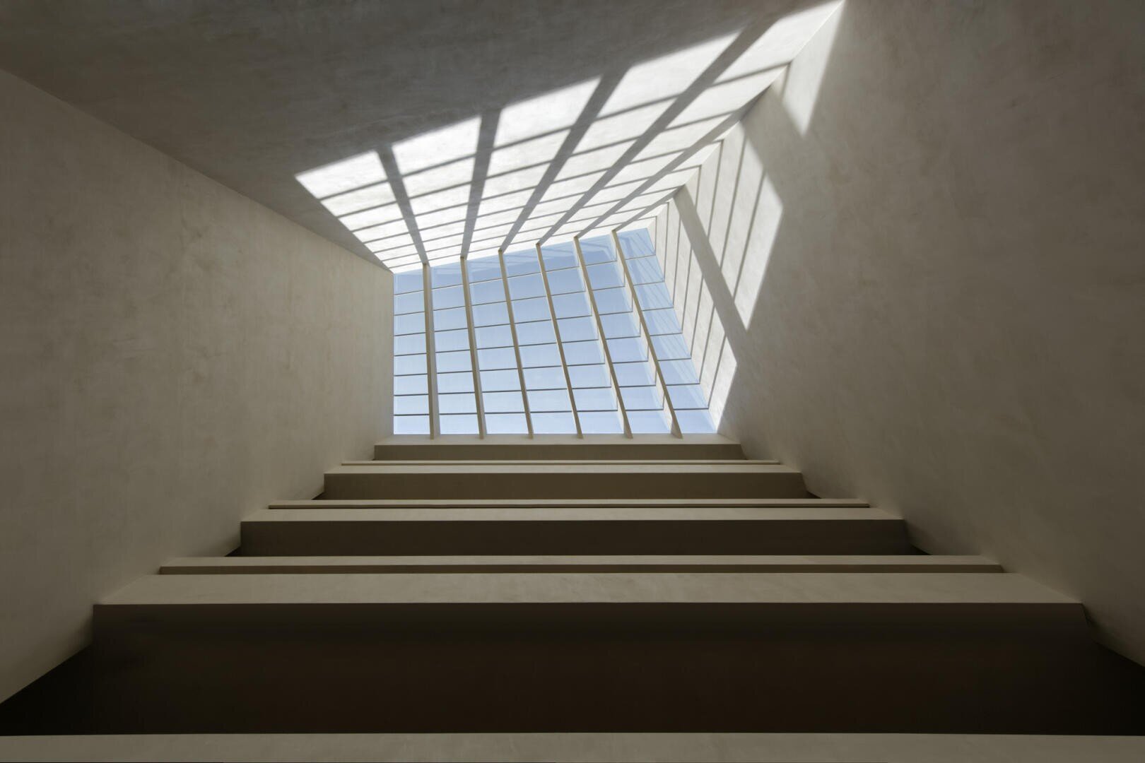 Der Blick über eine minimalistische Treppe auf ein großes, schräges Oberlicht mit Gittermuster. Sonnenlicht strömt hindurch und wirft geometrische Schatten auf Wände und Stufen. Die Umgebung besticht durch einen klaren, modernen Architekturstil.