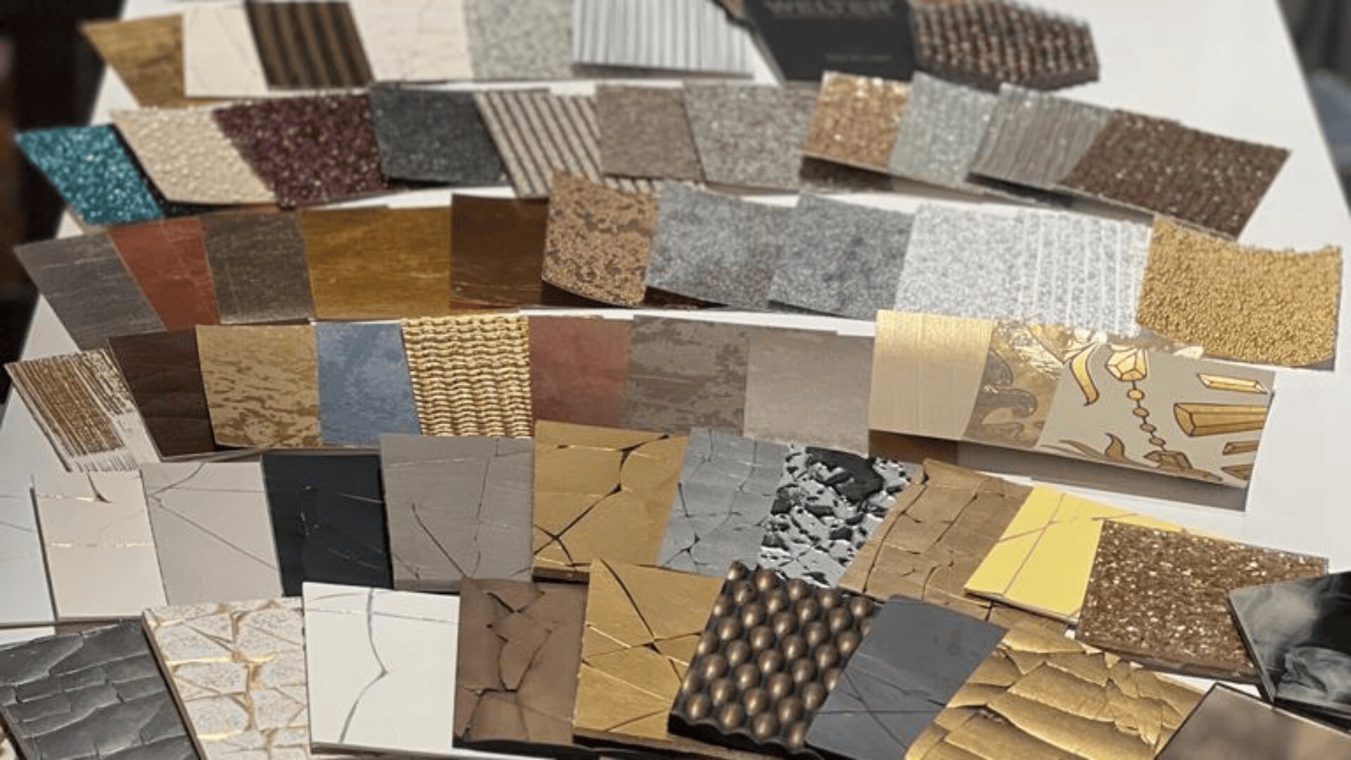 A variety of rectangular material samples are arranged on a table, showcasing different colors, textures, and patterns, including metallic, marble, glitter, woven, and stone-like finishes.