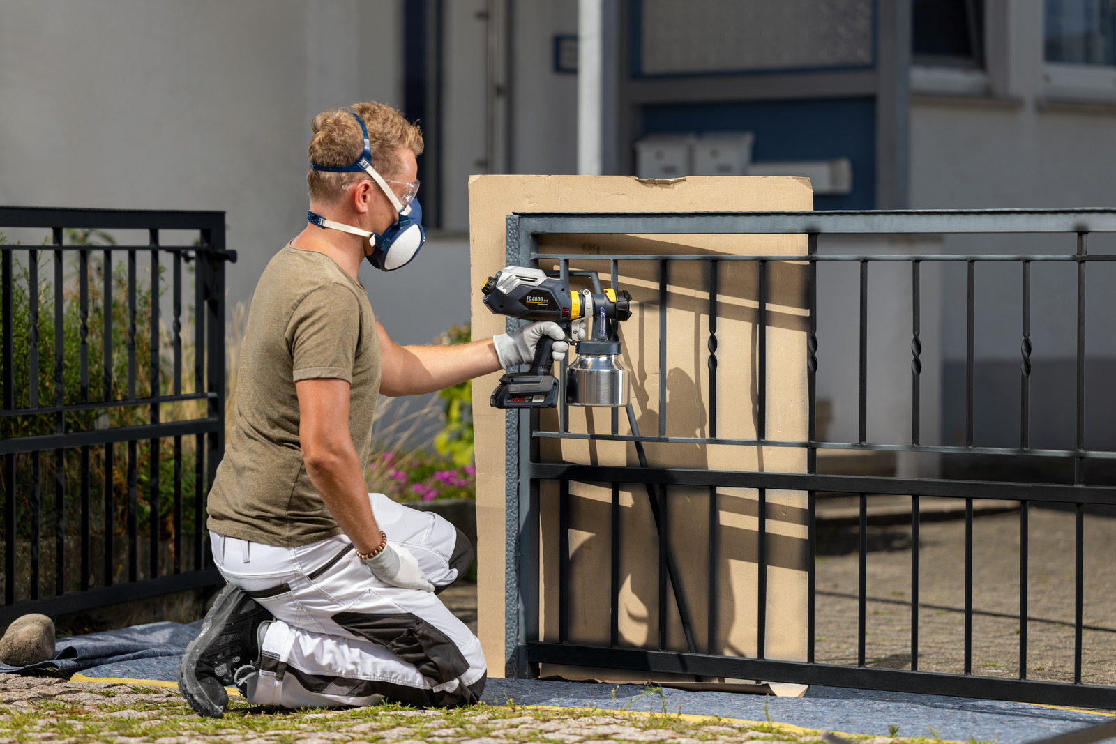 A person wearing a respirator mask, gloves, and kneepads uses a paint sprayer to paint a black metal fence outdoors, with a tarp covering the ground underneath the work area.