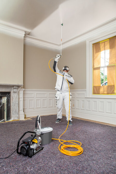 A person in protective gear uses a paint sprayer to paint a white ceiling in a room, with windows and fireplace covered for protection and equipment on the carpeted floor.