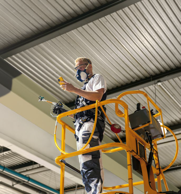 Ein Arbeiter in Schutzkleidung und Atemschutzmaske steht auf einer gelben Scherenbühne und überprüft mit einem Handgerät die Ausrüstung in der Nähe einer Metalldecke in einem Industriegebäude.