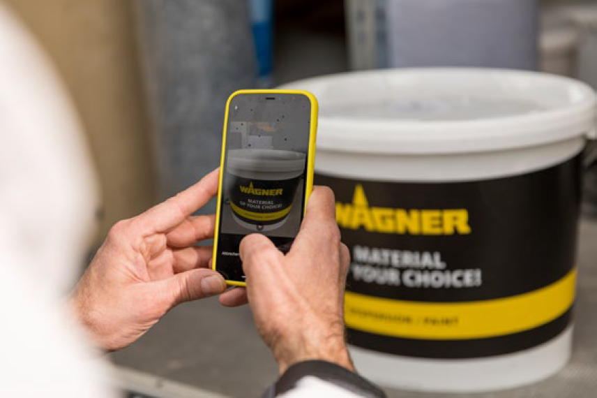 Eine Person hält ein Smartphone und fotografiert einen großen weiß-schwarzen Farbeimer mit der Aufschrift „Wagner“. Auf dem Display ist das Bild desselben Eimers zu sehen.