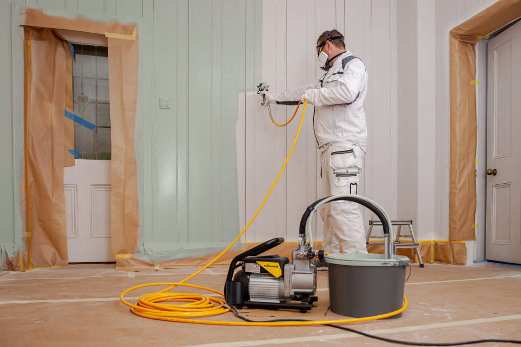 A person in protective clothing and mask uses a paint sprayer to paint white walls in a room. Doors and trim are covered with paper and tape, and a paint sprayer with a yellow hose is on the floor.